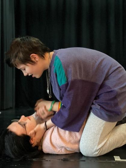 Zwei Mädchen* üben eine Theaterszene. Eine Person liegt auf dem Boden, die andere beugt sich über sie und hält ihren Kopf.