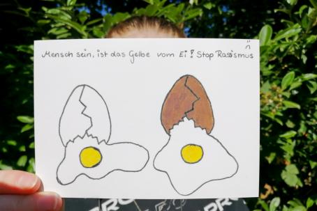 Eine Person hält eine Postkarte hoch, ein braunes und ein weißes Ei, beide sind zerbrochen, das Innere sieht gleich aus